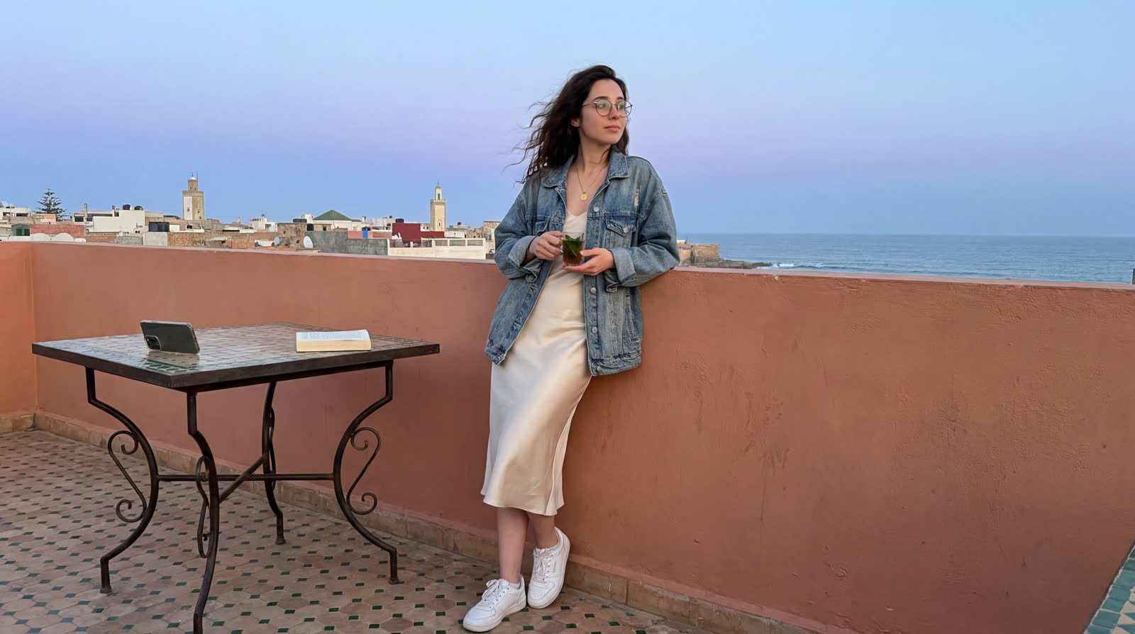 August on a riad rooftop in the Essaouira medina before dawn, holding a glass of mint tea, the Atlantic beyond the rooftops
