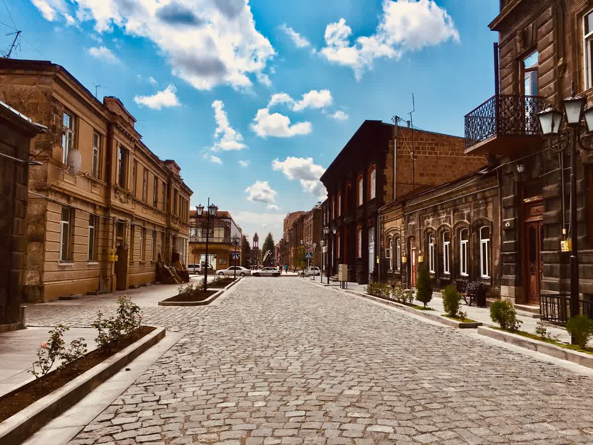 Abovyan Street in Gyumri's Kumayri historic district, cobblestone road flanked by two-story stone buildings in warm brown and ochre tones with arched windows and wrought-iron balconies